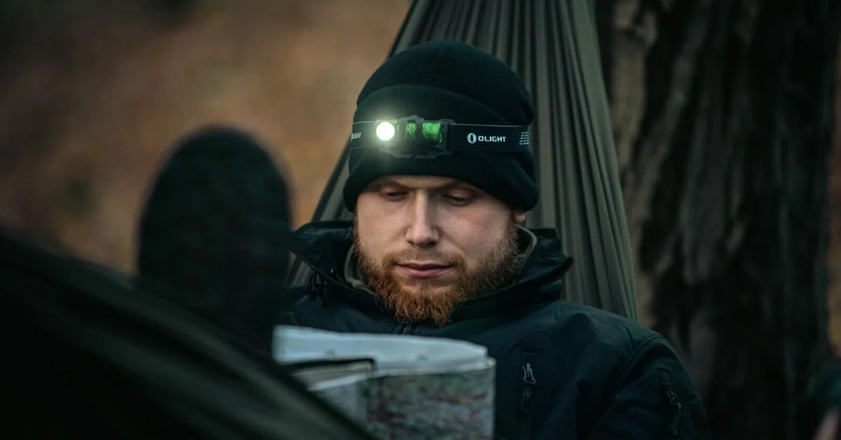Man in a beanie and headlamp reading a map beside a hammock at dusk